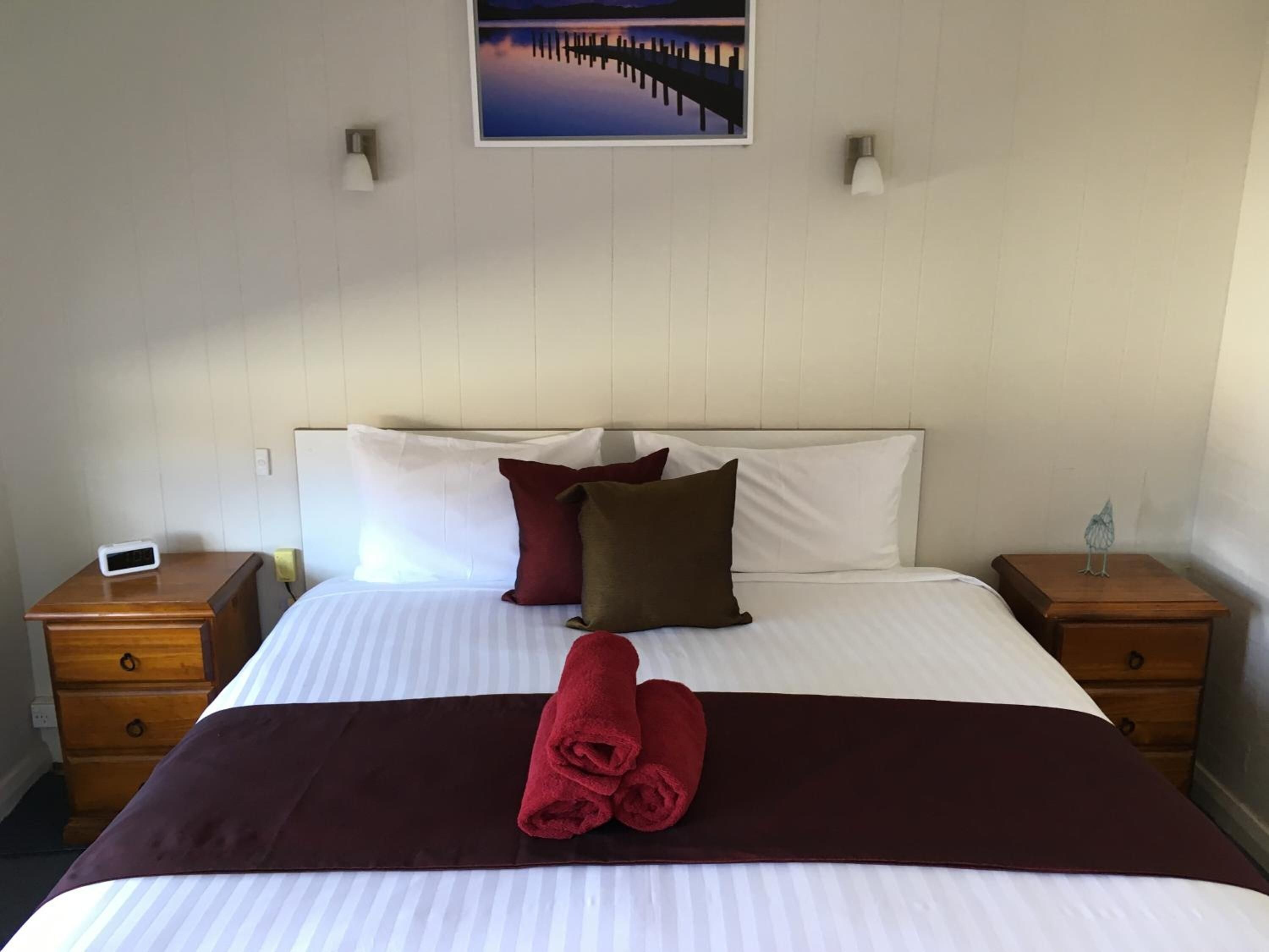 A hotel room with a bed, pillows, a folded towel, a nightstand, a wall-mounted lamp, and a painting above the bed.