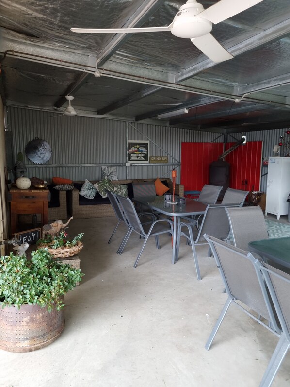 Un área de estar al aire libre techada con sillas de metal y una mesa, un ventilador de techo y una pared roja con un cartel que dice 'DRINK UP'.