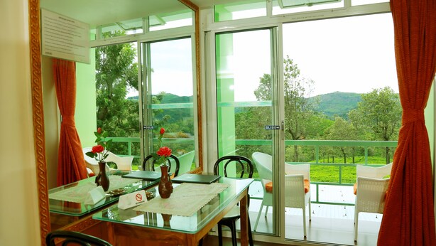 Un comedor con mesa de vidrio, sillas, un florero con flores y vistas a zonas verdes y colinas a través de amplios ventanales.