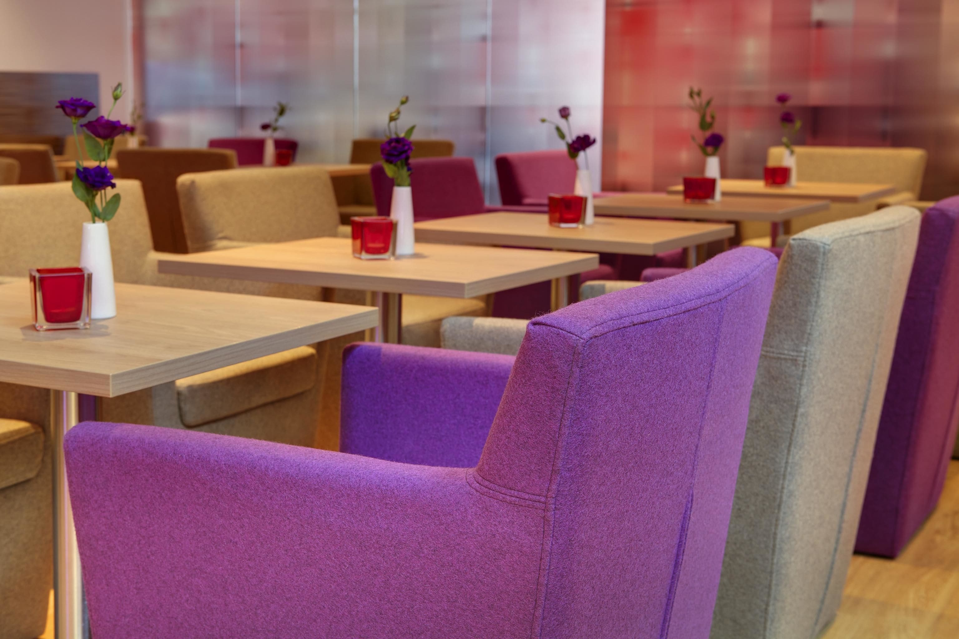 Un restaurante con sillas tapizadas en tonos morado y gris, mesas de madera y candelabros rojos con flores moradas.
