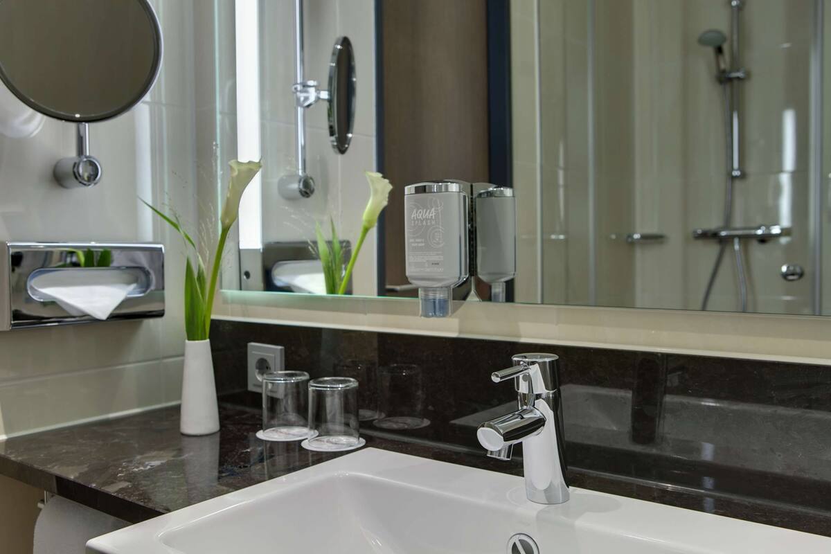 Een moderne badkamer met een witte wastafel, chromen kraan en een grote spiegel. Op het aanrecht staan twee glazen potten, en een vaas met groene planten.
