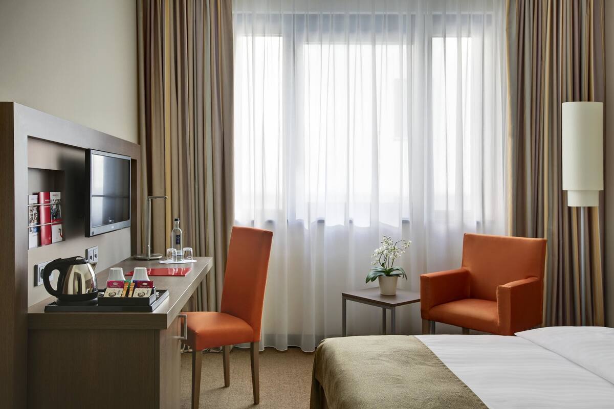 Een hotelkamer met een bed, twee oranje fauteuils, een bureau met een televisie, een kleine tafel met een plant en een raam met gordijnen.