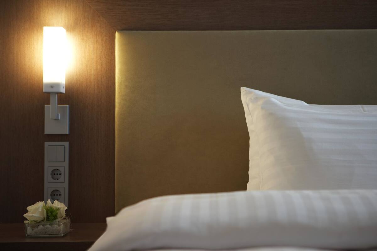 Een keurig opgemaakt bed met een wit kussen en hoofdbord, een nachtkastje met een lamp, en een kleine vaas met bloemen.