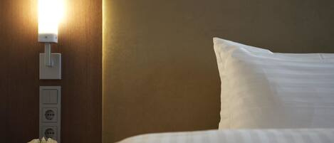 A neatly made bed with a white pillow and a headboard, a bedside table with a lamp, and a small vase with flowers.