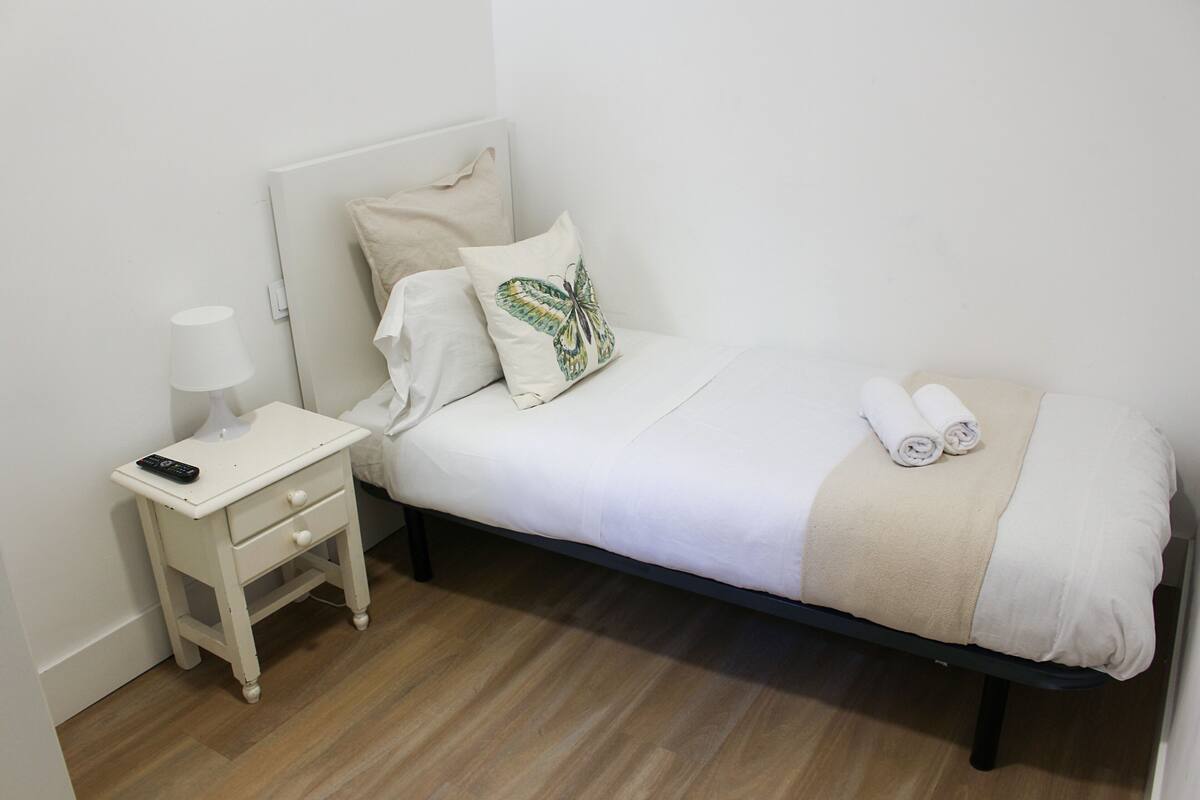 A neatly made bed with a white and beige blanket, a pillow with a butterfly design, and rolled towels on the bed.