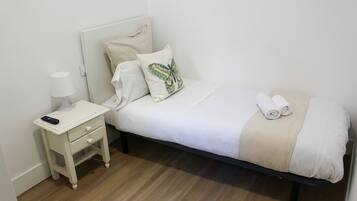 Una cama bien tendida con ropa blanca y un plaid color beige, una mesita de noche con una lámpara y un control remoto sobre la mesa.