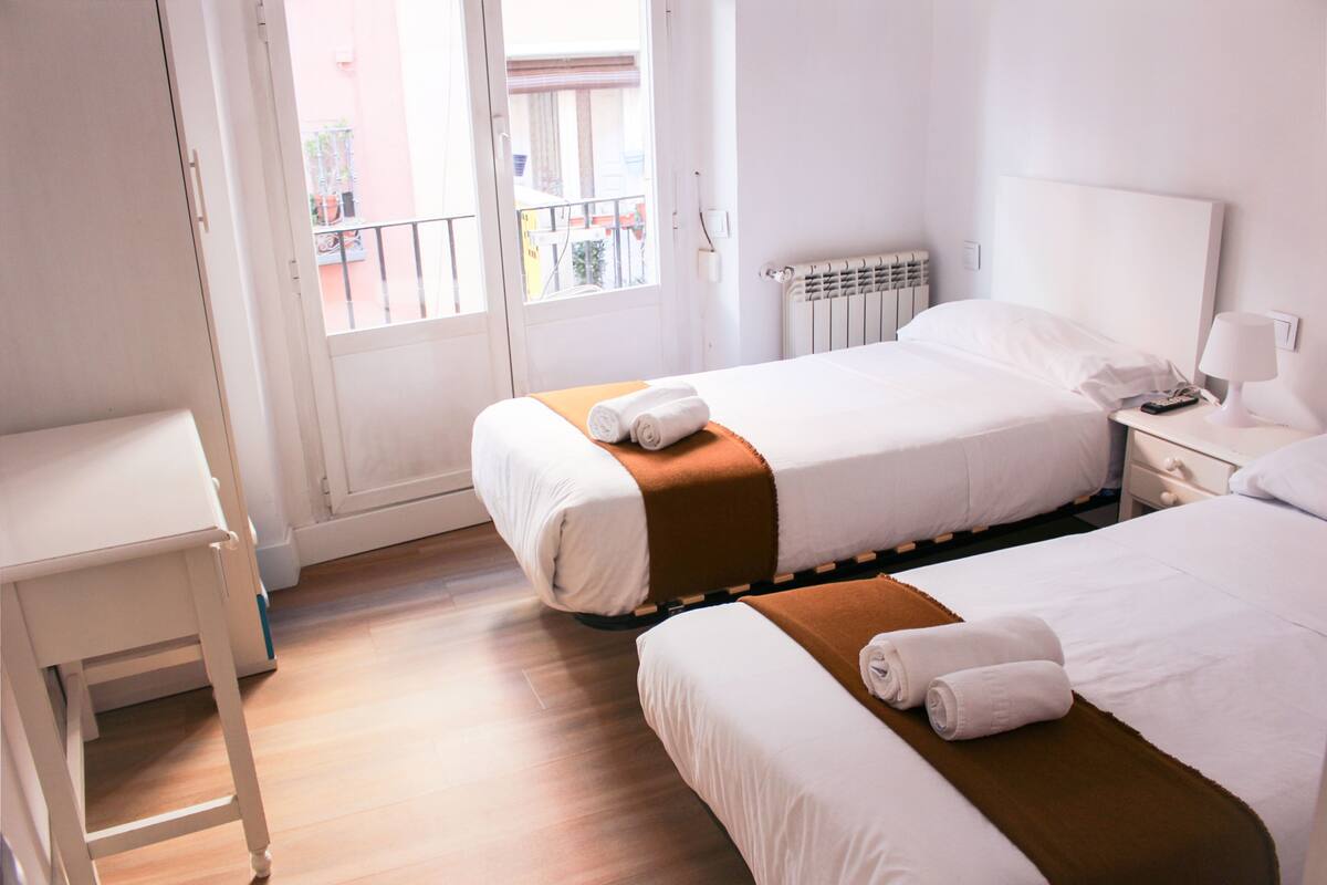 A hotel room with two beds, a small table, a nightstand, and a window with a view of the outside.