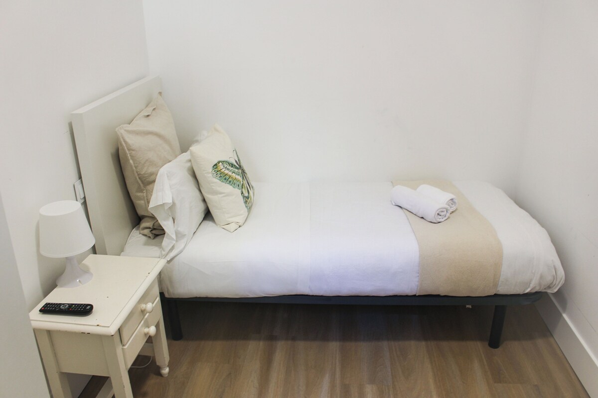 Una cama pequeña y ordenada con una colcha blanca y beige, una toalla enrollada y un control remoto sobre la mesita de noche.