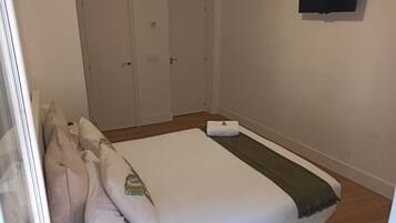 Un dormitorio con cama, almohadas, una colcha, un televisor en la pared y dos puertas.