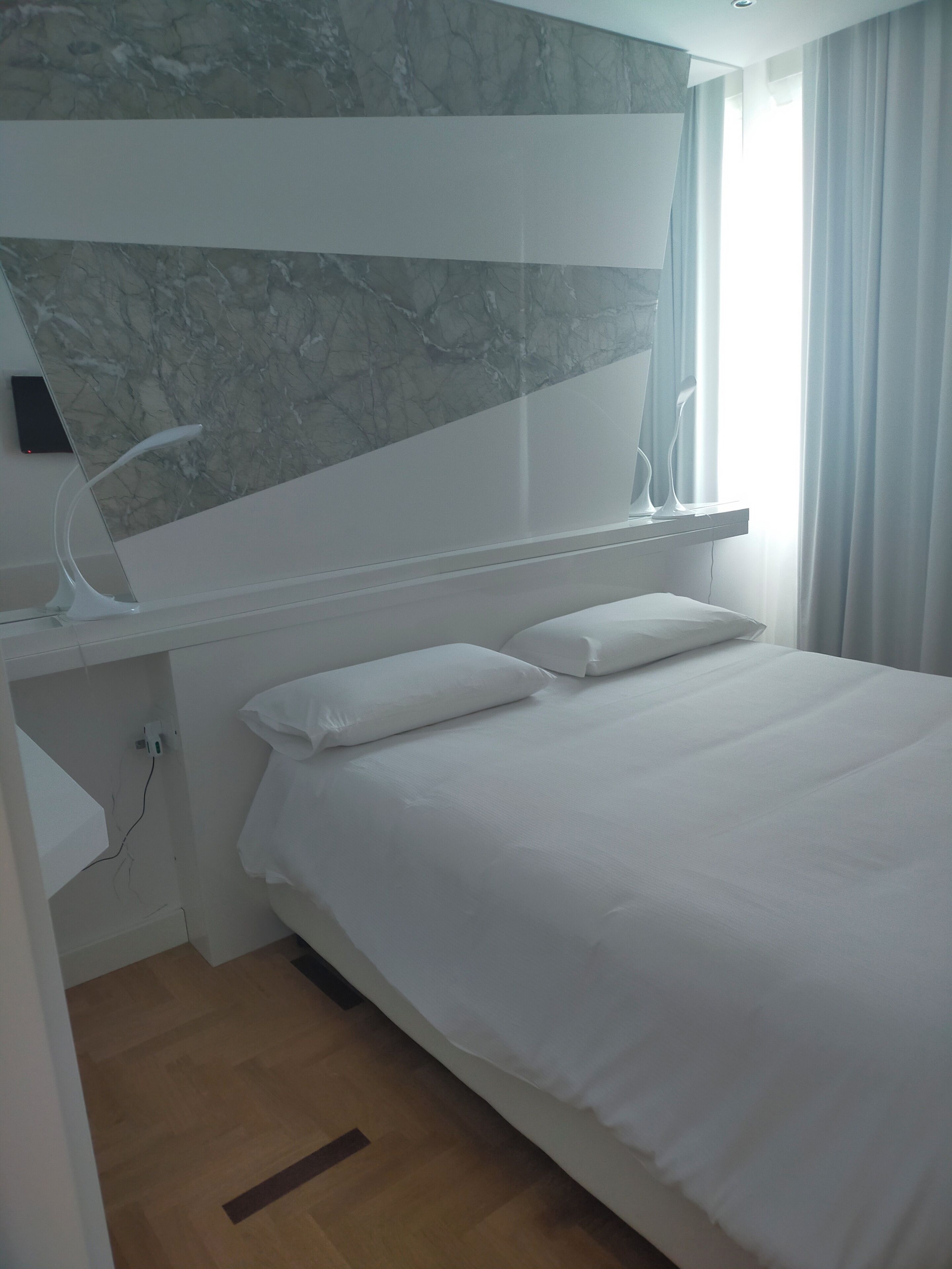 Ein modernes Hotelzimmer mit einem großen Bett, einer Marmorwand, einem Nachttisch mit Lampe und Blick ins Freie.
