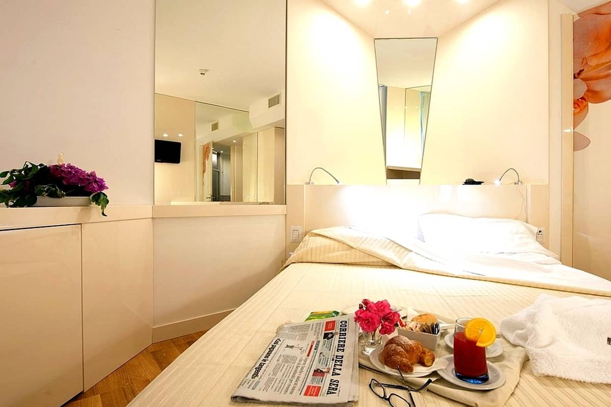 Una habitación de hotel con una cama, una mesita de noche con un periódico, un vaso de bebida roja, un croissant y una toalla.