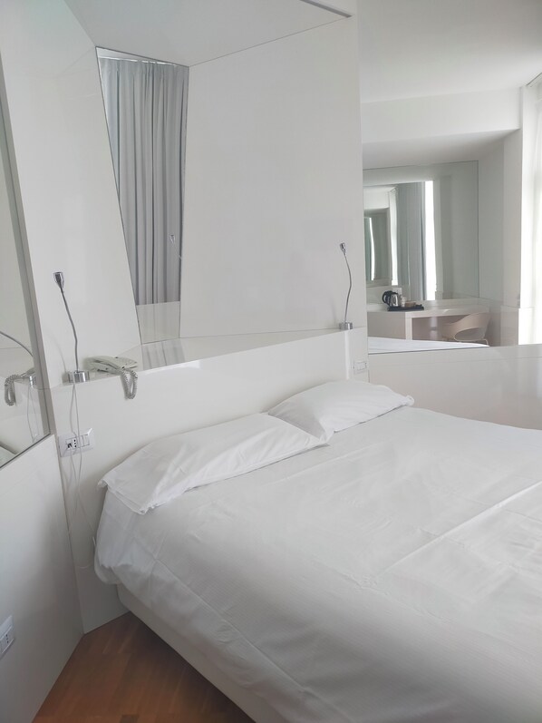 A clean, modern hotel room with a large bed, a bedside table, a mirror, and a bathroom visible in the background.
