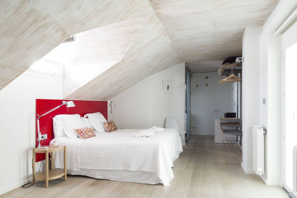 Une chambre avec un plafond en pente, un lit avec une literie blanche, une tête de lit rouge et une table de chevet avec une lampe.