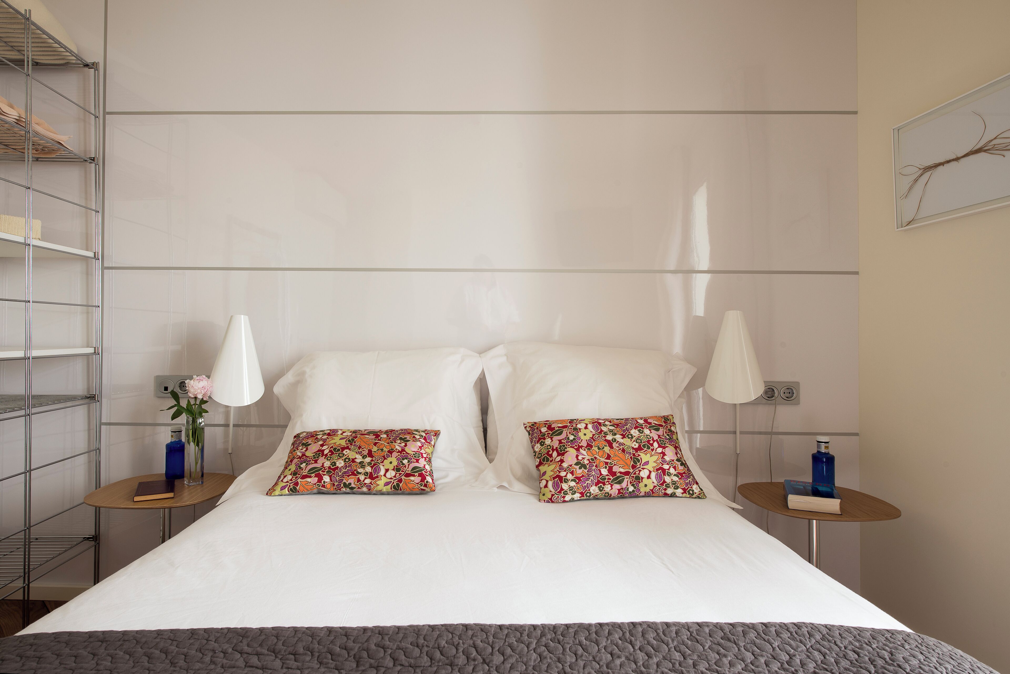 A neatly made bed with two decorative pillows, a bedside table with a book and a vase, and a wall-mounted shelf with a vase.