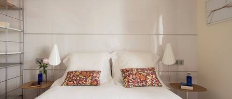 A neatly made bed with two decorative pillows, a bedside table with a book and a vase, and a wall-mounted shelf with a vase.