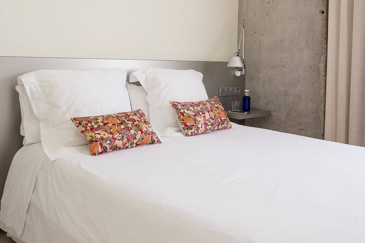 Un lit bien fait, avec du linge de lit blanc et deux coussins décoratifs, une table de chevet avec une lampe et une bouteille, et un mur sobre avec une fenêtre.