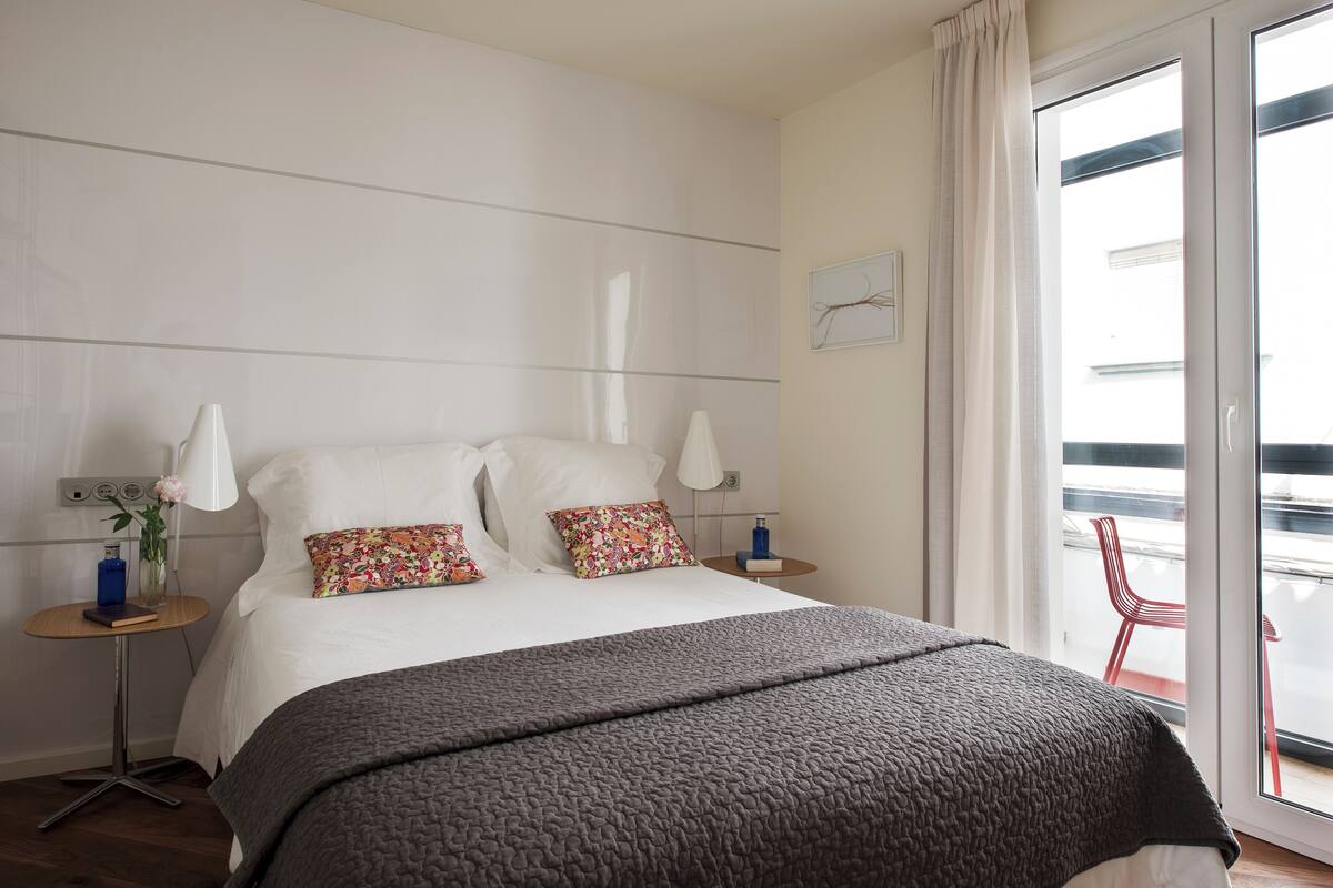 Une chambre avec un grand lit, deux tables de chevet, une petite table et une vue sur le balcon extérieur.