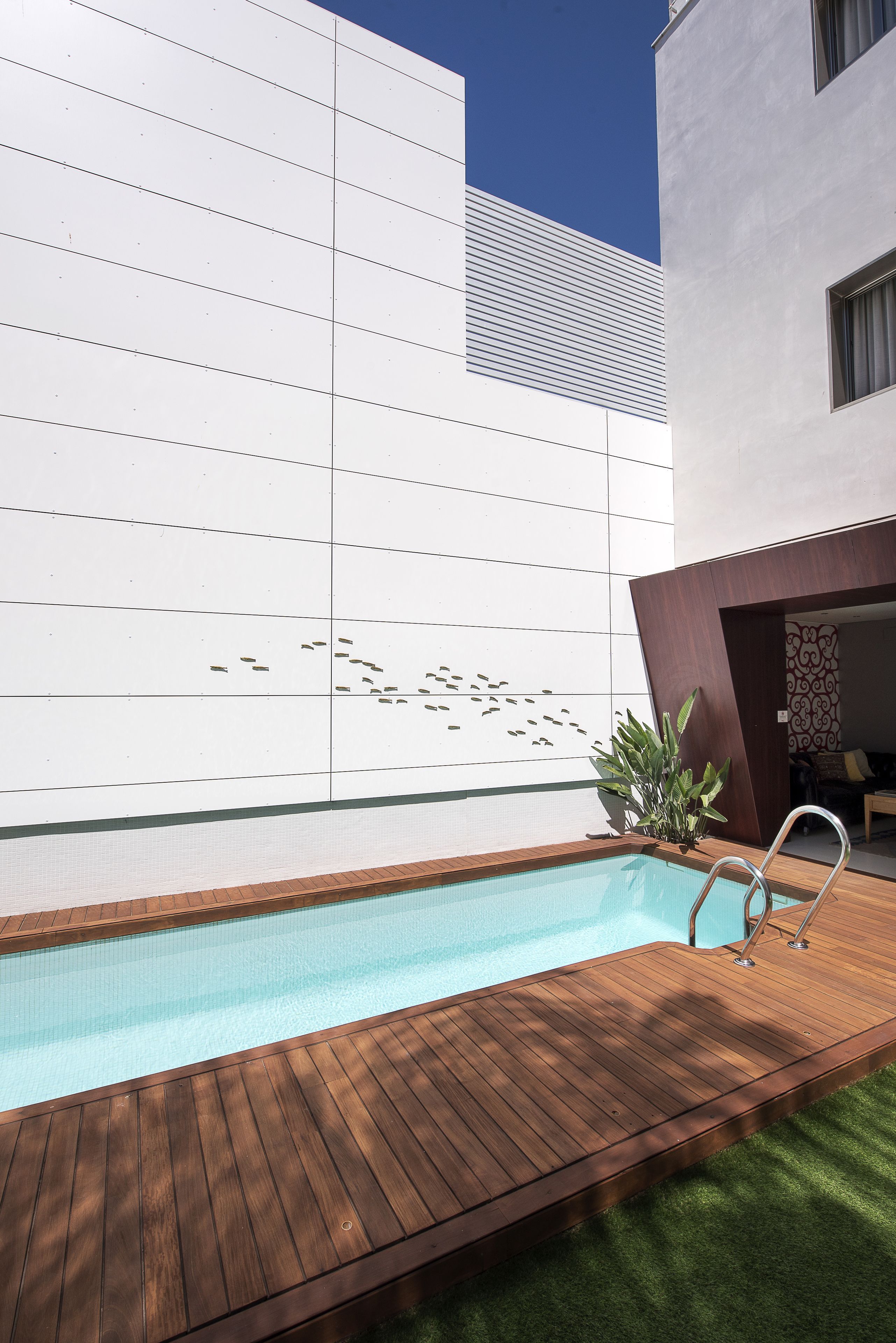 A swimming pool with a wooden deck, a white wall with a geometric pattern, and a door leading to an interior space.