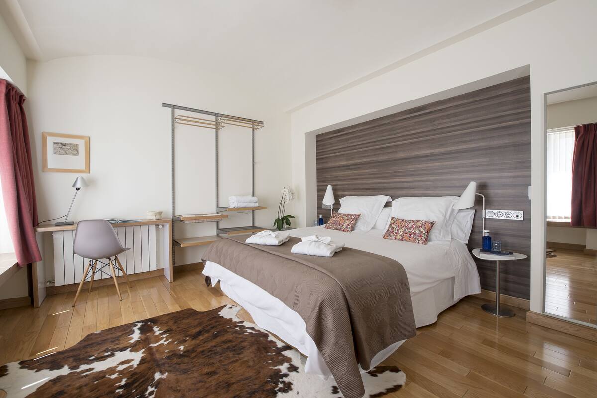 Une chambre moderne avec un plancher en bois, un grand lit doté d’une tête de lit marron, un bureau avec une chaise et une petite table ronde avec une lampe.