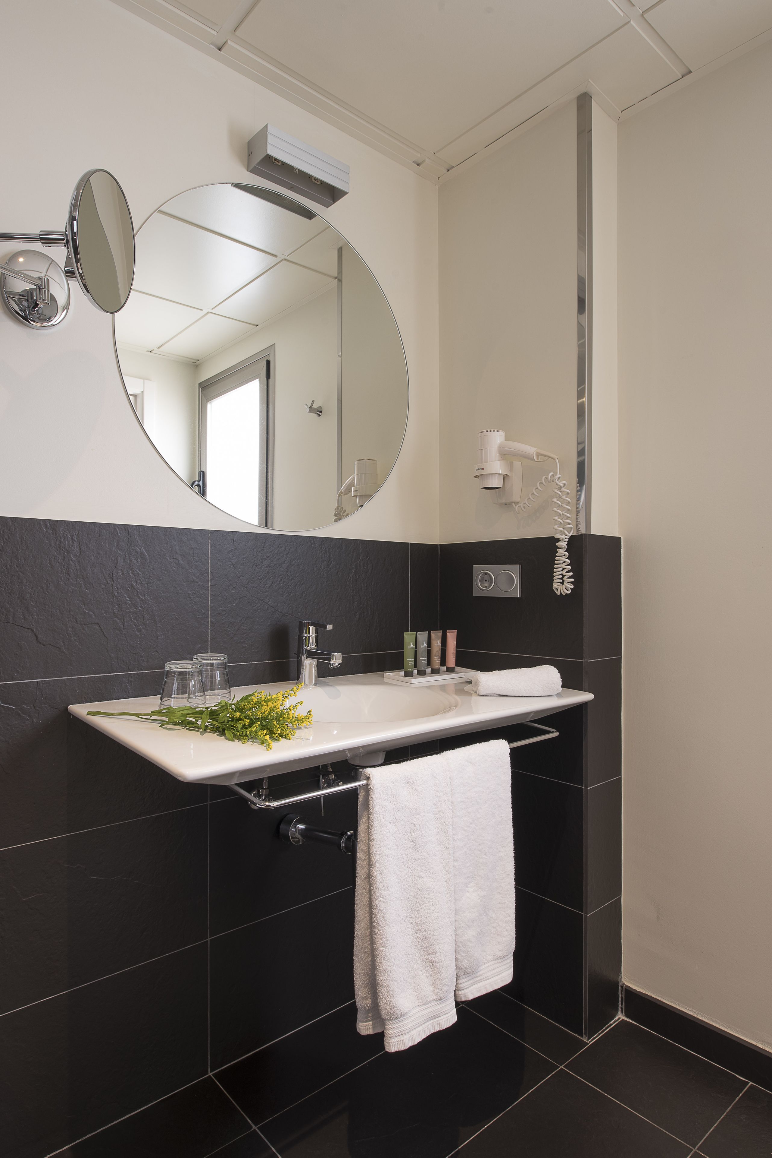 A modern bathroom with a white sink, black tiles, a large mirror, a handheld showerhead, and a towel rack.