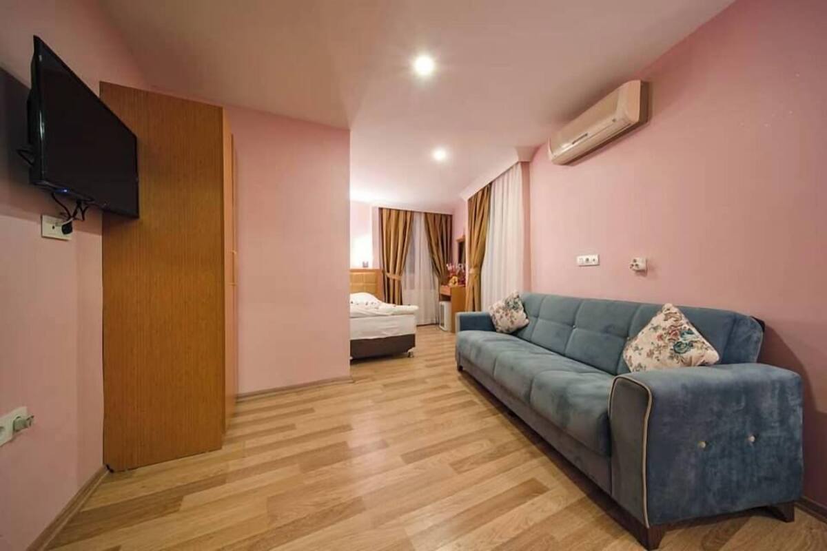 A room with a wooden floor, a blue sofa, a television mounted on the wall, and a bed in the background.