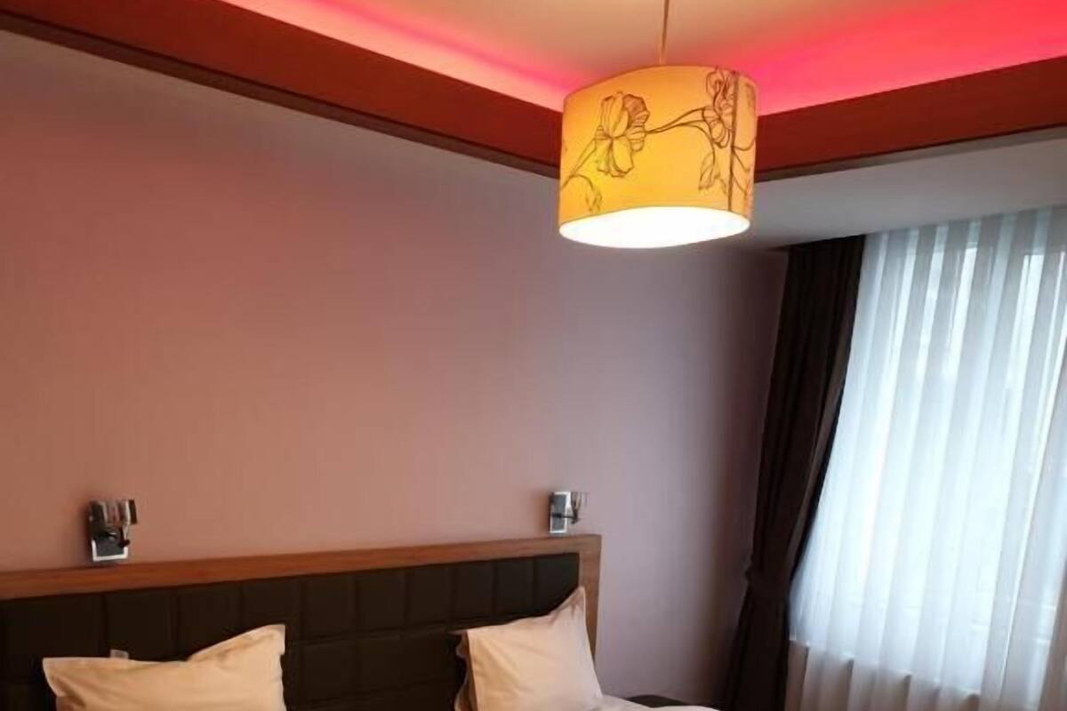 A hotel room with two beds, each with a towel arranged in a swan shape. There is a ceiling light fixture above the beds, and a window with curtains.