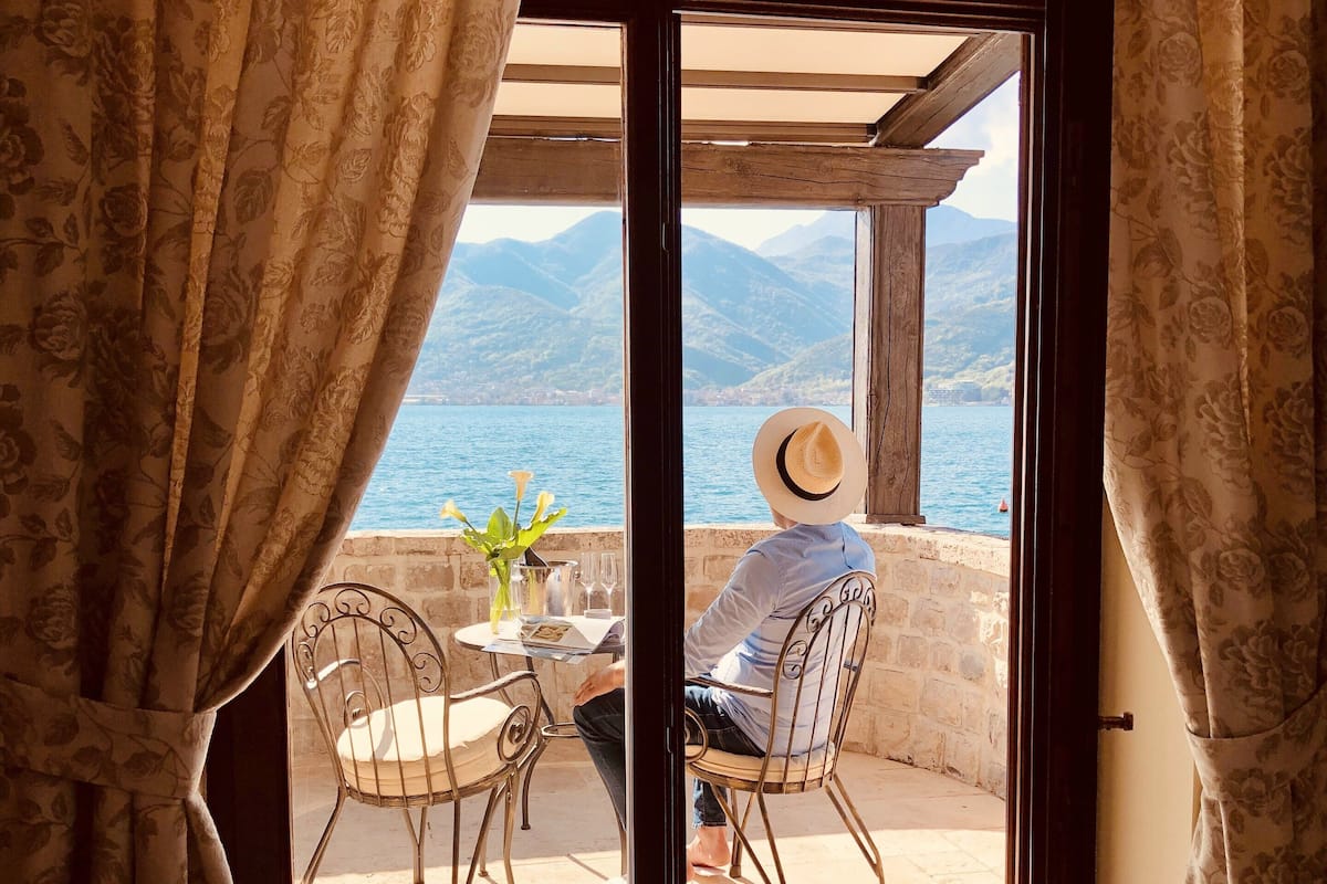 A room with a view of a body of water and mountains, featuring a table with chairs, a hat, and a vase with flowers.