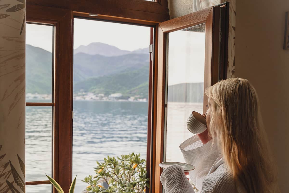 A person standing by a window with a view of a body of water and mountains.