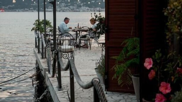 Un restaurante frente al agua con terraza, un camino de piedra y vistas al mar y las montañas.