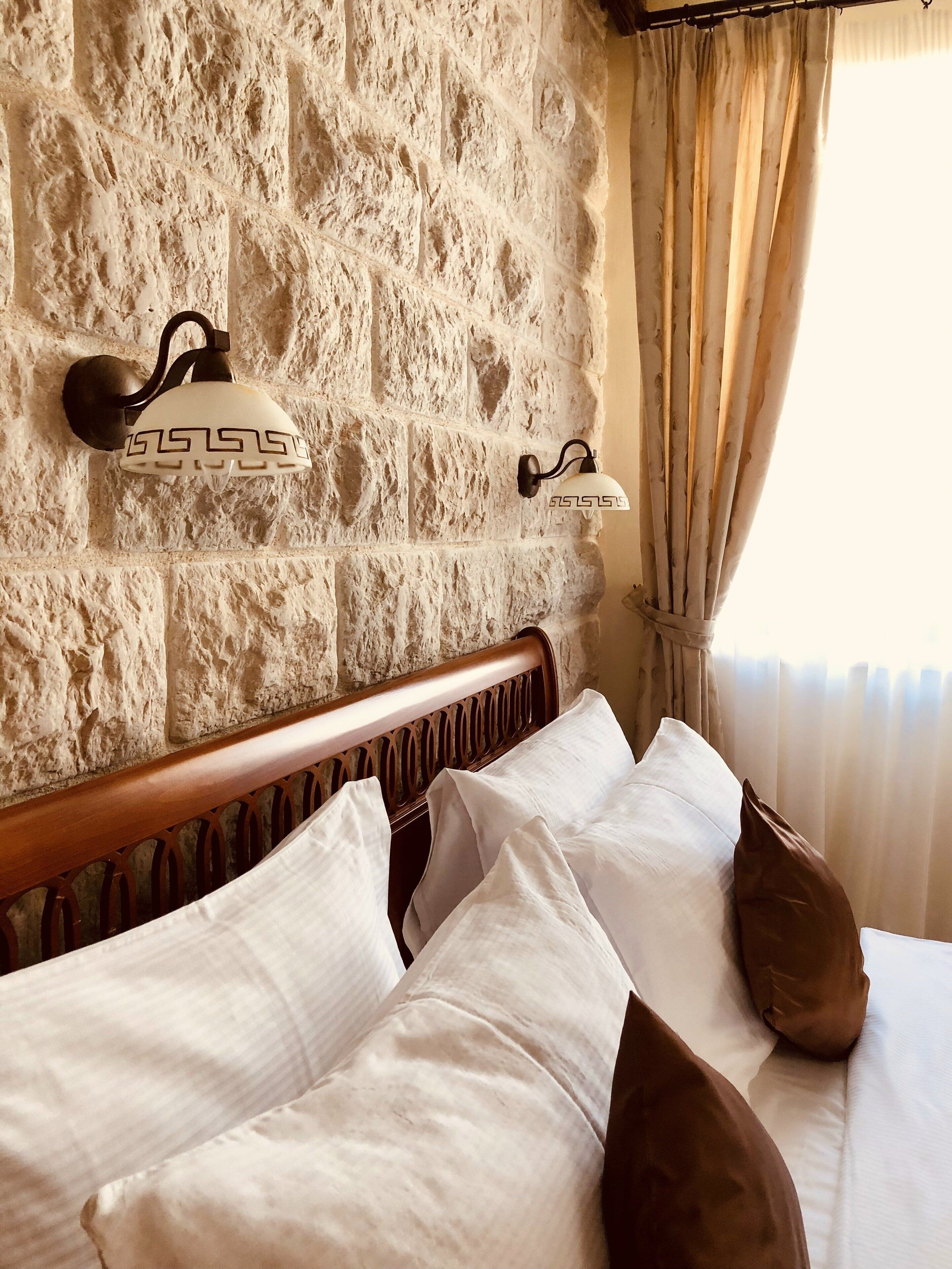 Una cama con almohadas blancas y marrones, un cabecero de madera y una pared de piedra con dos luces de pared.