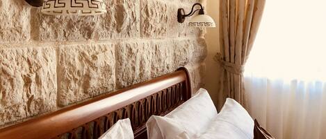Una cama con almohadas blancas y marrones, un cabecero de madera y una pared de piedra con dos luces de pared.