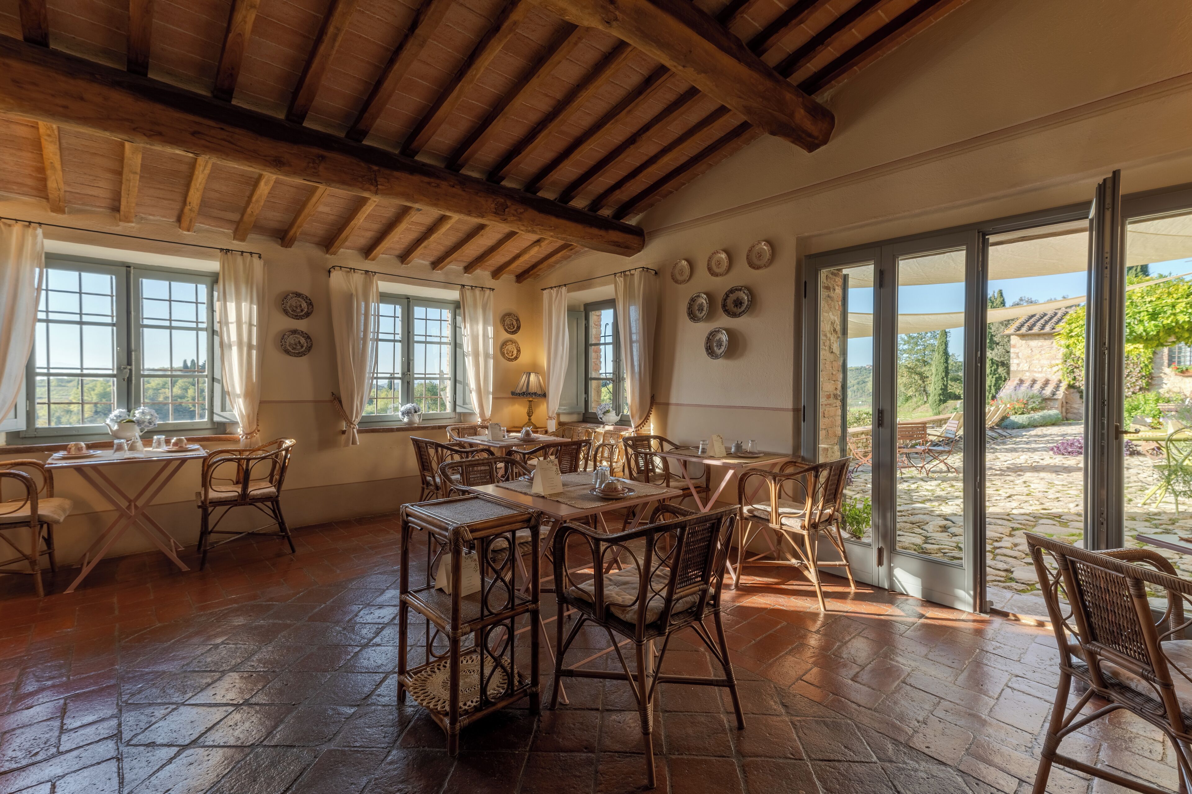 A spacious dining area with wooden tables and wicker chairs, large windows with curtains, and a view of the garden.