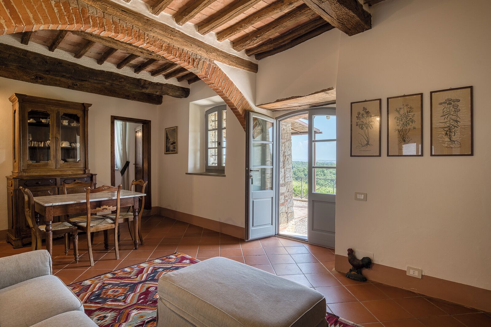 Un comedor con una mesa y sillas de madera, un armario con puertas de vidrio y vistas a un balcón exterior desde un ventanal abierto.