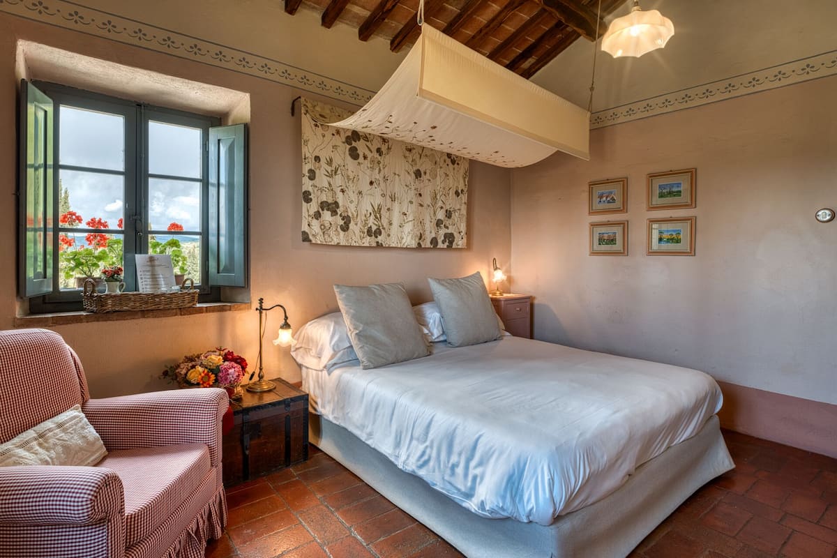 A bedroom with a bed, a chair, a nightstand, a lamp, a window with flowers, and a wall with framed pictures.