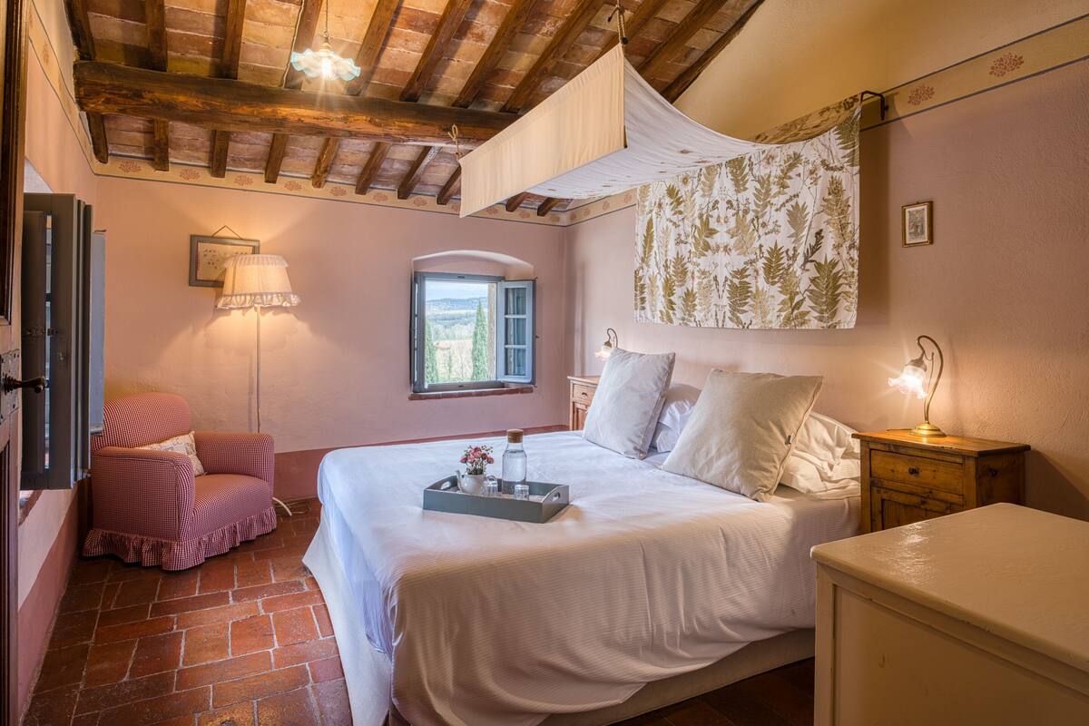 A bedroom with a large bed, a chair, a nightstand, a lamp, and a window with a view.