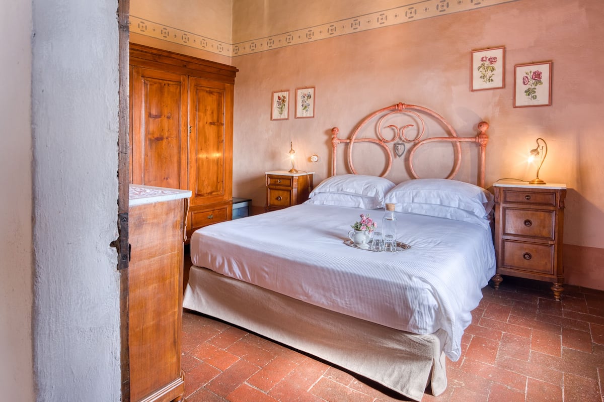 A bedroom with a large wooden wardrobe, a bed with white linens, two bedside tables with lamps, and floral wall art.