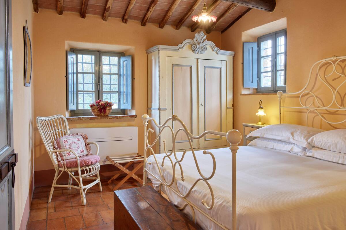 A bedroom with a bed, a chair, a wardrobe, and a window with a basket of flowers.