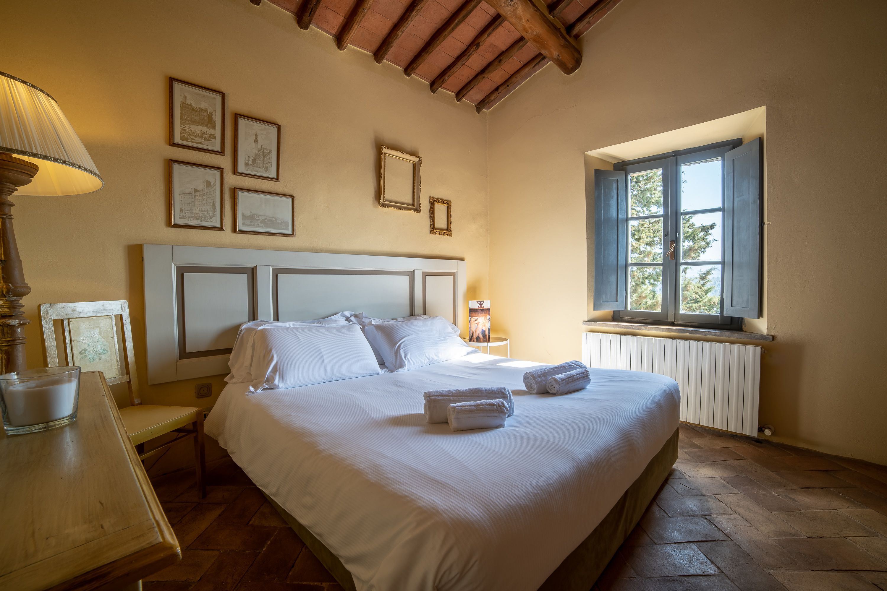 A neatly made bed with white linens, a bedside table with a lamp, and a window with a view of trees.
