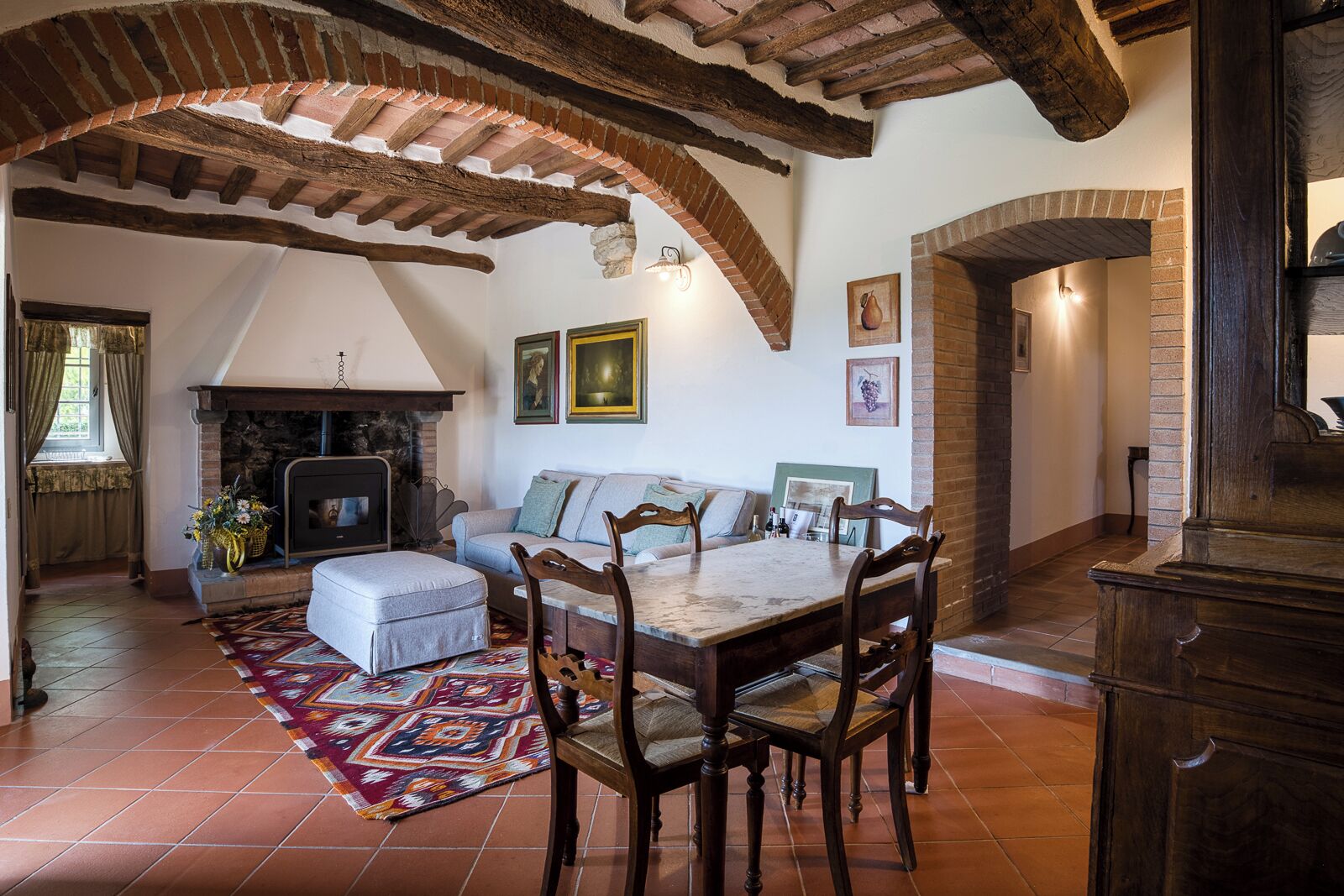 A rustic living room with exposed wooden beams, a fireplace, a dining table with chairs, and a patterned rug.