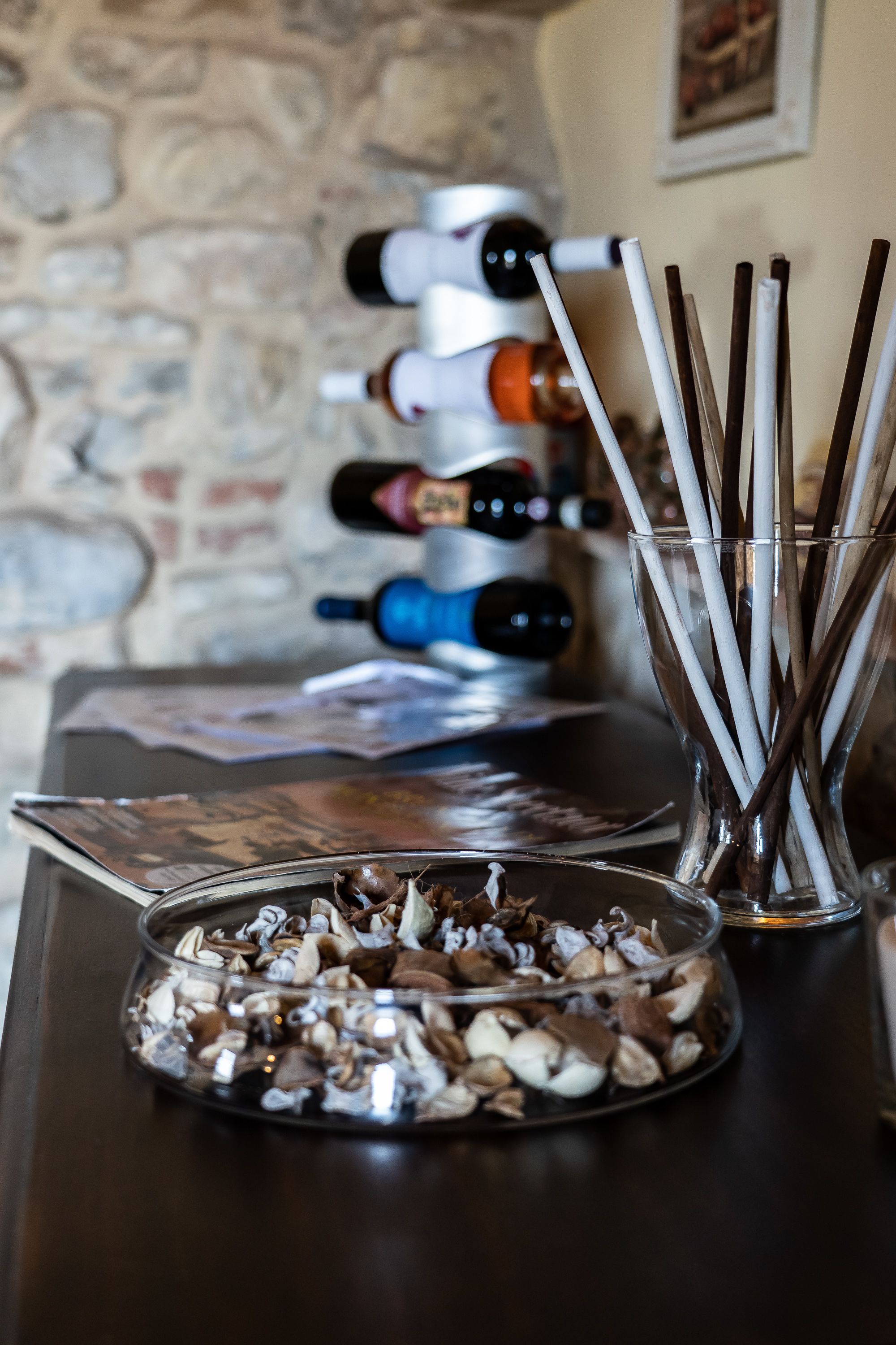 A table with a glass bowl of nuts, a glass with straws, and a row of wine bottles.