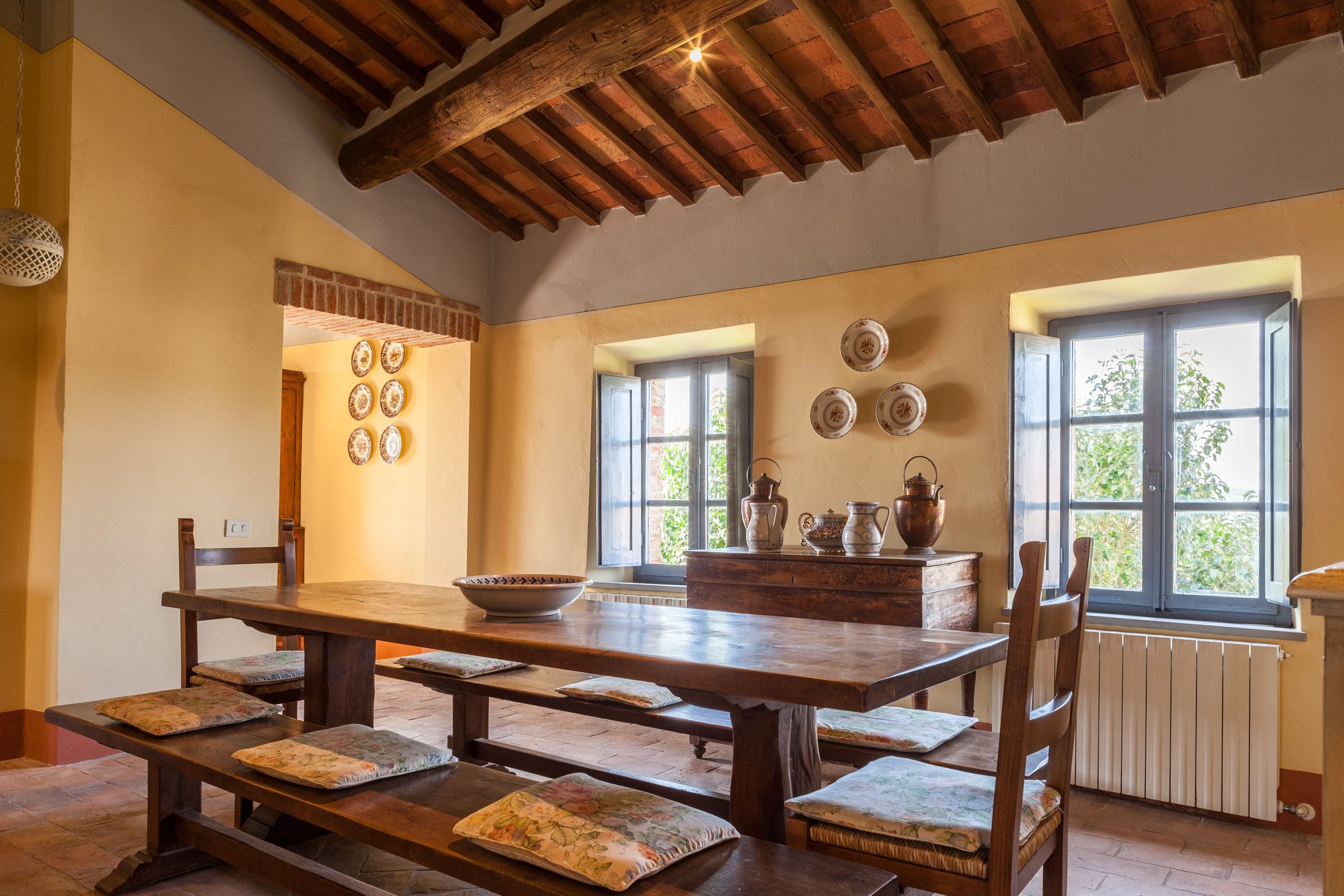 A dining area with a wooden table and benches, a brick wall, and a window with a view.