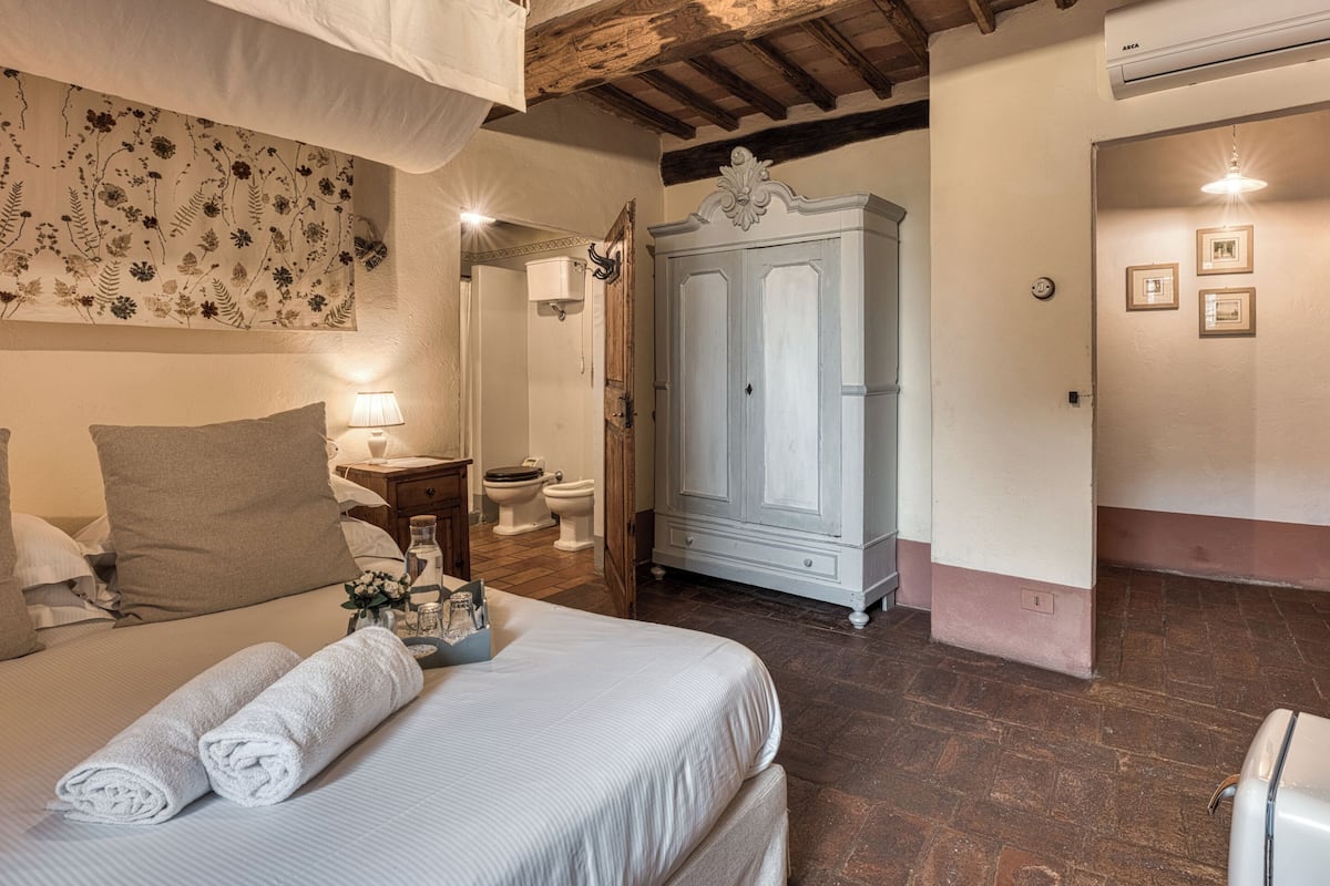 A bedroom with a bed, a nightstand, a mirror, a wardrobe, and a bathroom visible in the background.