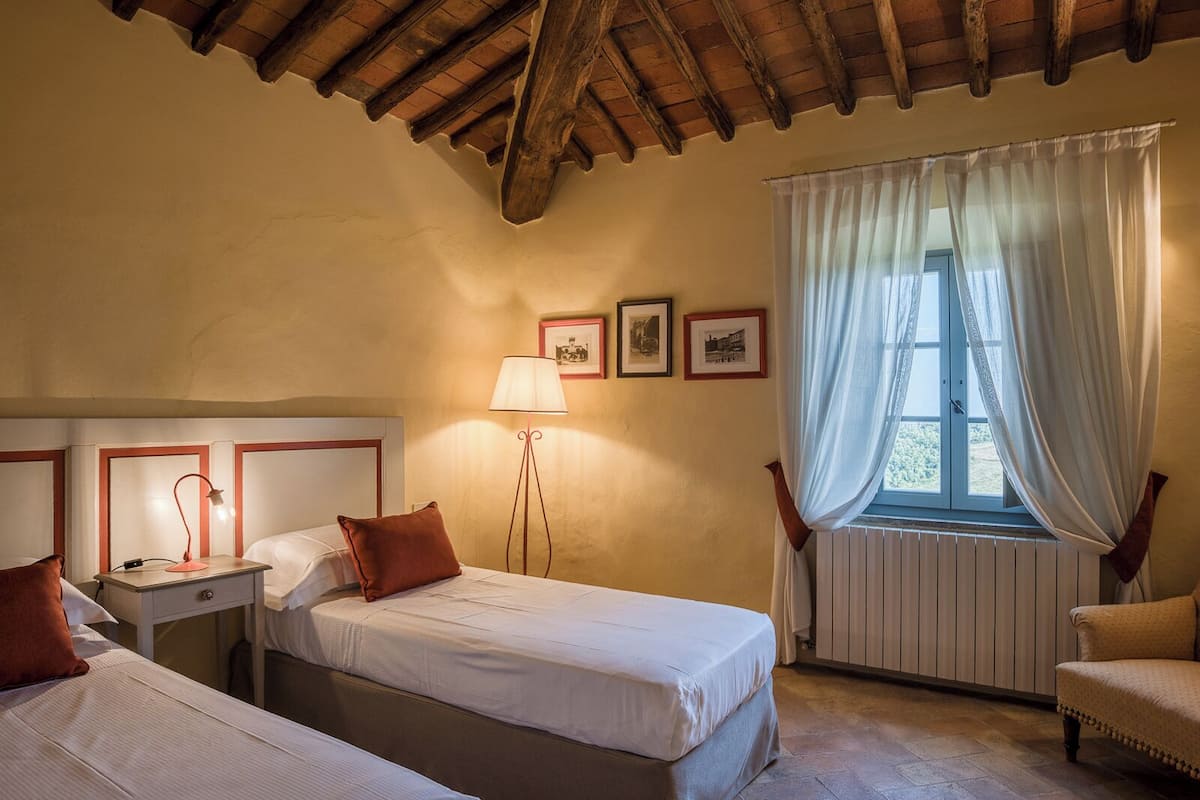 A hotel room with two beds, a nightstand, a lamp, a chair, and a window with curtains.