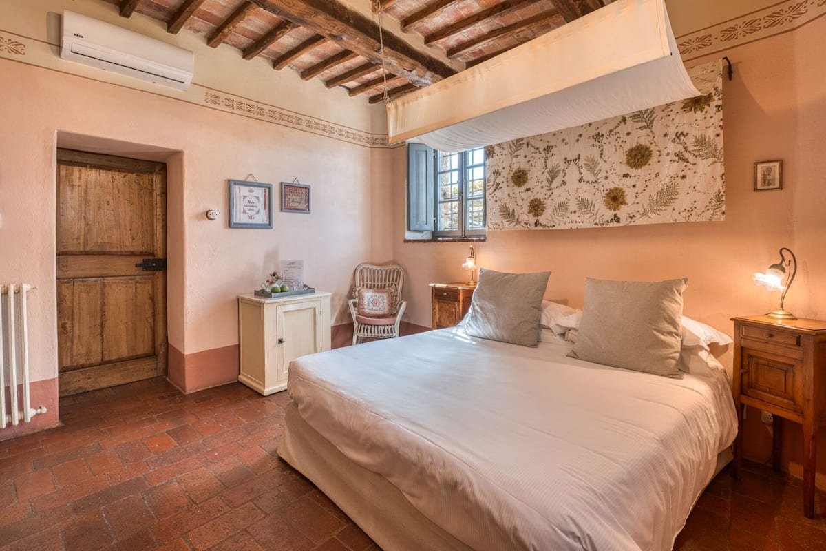 A bedroom with a large bed, a wooden door, a small cabinet, a chair, a nightstand with a lamp, and a window with floral wallpaper.