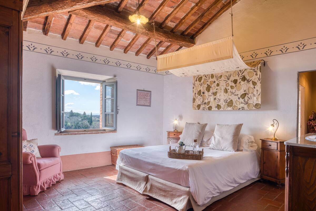 A bedroom with a bed, a window, a wooden ceiling, and a wall with a patterned tapestry.