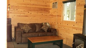 A wooden cabin interior with a brown sofa, patterned cushions, a wooden coffee table, a fireplace, and a window with a view of trees.