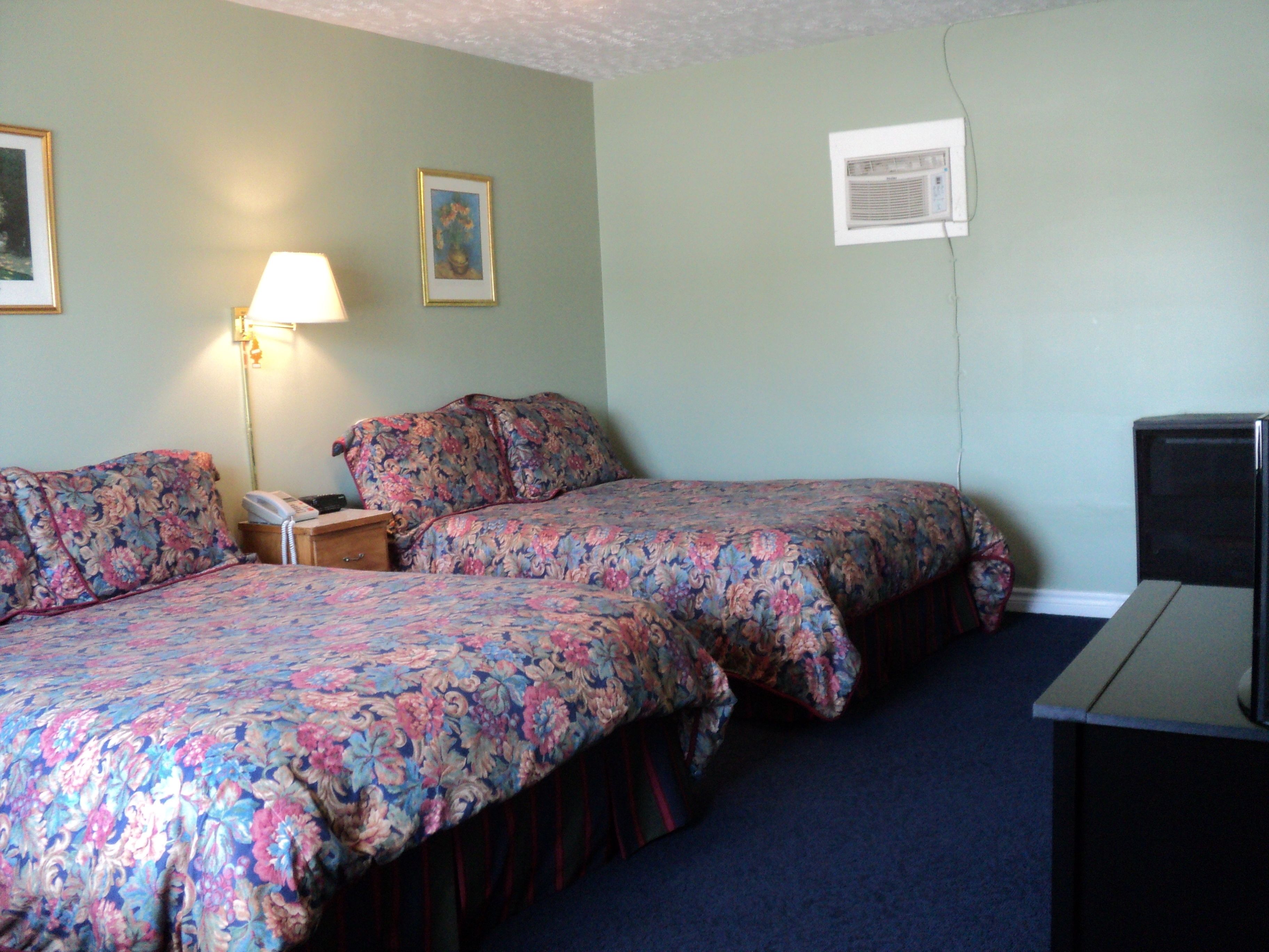 A hotel room with two beds, a bedside table with a lamp, a framed picture on the wall, and a television.
