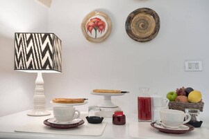 Una mesa de desayuno con mantel blanco, una lámpara estampada, una cesta de frutas y una jarra roja para bebidas.