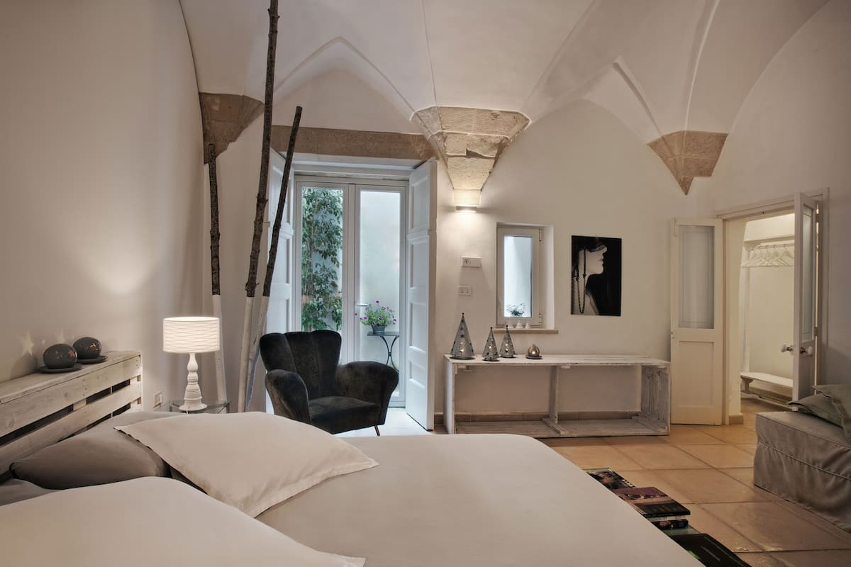 A bedroom with a large bed, a chair, a console table with decorative items, a mirror, and a door leading to a bathroom.