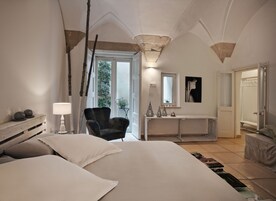 Quarto com uma cama grande, uma cadeira, uma mesa consola com objetos decorativos, um espelho e uma porta que dá para uma casa de banho.